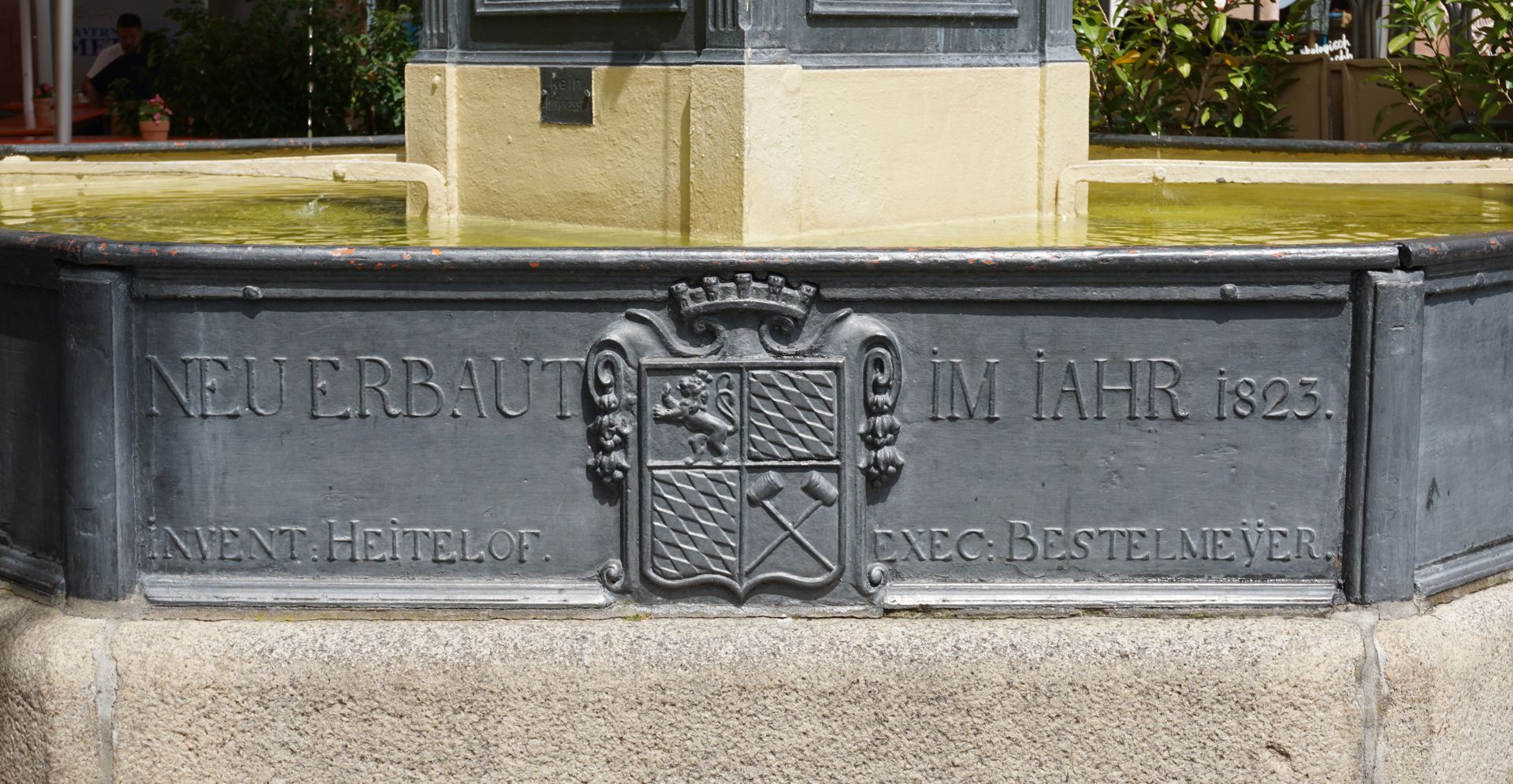 Pferdebrunnen Wasserbecken, Südseite mit Inschrift: "NEU ERBAUT IM Jahr 1823. INVENT(OR) (=Erfinder) HEITELOF. EXEC(UTOR) (=Ausführender) BESTELMEYER" / Stadtwappen von 1808 mit bayerischen Rauten, dem Pfälzer Löwen und zwei gekreuzten Bierschöpfen.